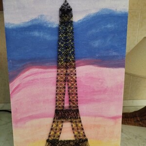 Può includere: Una rappresentazione della Torre Eiffel in filo nero su uno sfondo dipinto di un tramonto con tonalità rosa, blu e gialle.