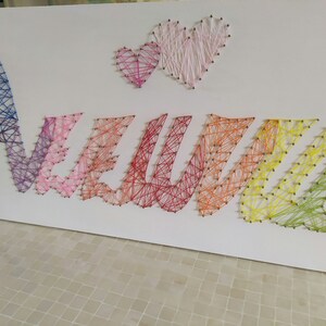 Può includere: Una tela bianca con la parola "NEVIL" scritta in arte di filo colorato. L'arte di filo è realizzata con fili di diversi colori avvolti attorno a chiodi martellati sulla tela. Due piccoli cuori si trovano nella parte superiore della tela, anch'essi realizzati in arte di filo.