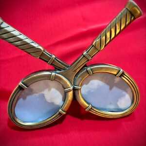 May include: A decorative, bronze-toned double picture frame. The frame features two oval openings, each designed to hold a photograph. The handles are textured with a woven pattern. The frame is set against a red background.