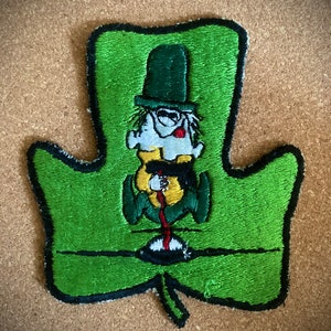 May include: Green felt patch in the shape of a maple leaf with a cartoon character wearing a green hat and holding a red stick.