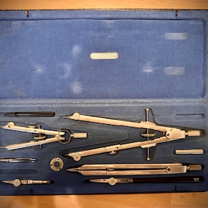 May include: A set of drafting compasses in a blue velvet case. The compasses are made of silver metal and have a variety of attachments. The case is labeled "GOMMUND".