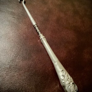 May include: A silver shoe horn with a decorative handle. The handle has a floral design and a small circle with a raised design.