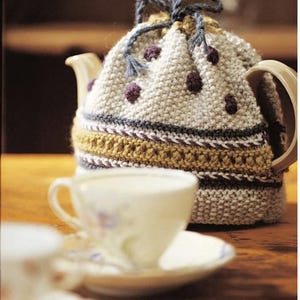 May include: A knitted teapot cozy with a brown, white, and gray striped pattern and purple polka dots. The cozy has a drawstring closure and is sitting on a wooden table next to a white teacup and saucer.