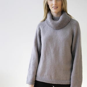 Op de afbeelding: Een lichtgrijze gebreide trui met een brede, geribbelde col. De trui met lange mouwen heeft een geribbelde zoom en manchetten. Het model draagt een zwarte broek. De trui is een comfortabele, casual stijl.