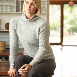 Könnte beinhalten: Eine Frau in einem hellgrauen Rollkragenpullover und einer schwarzen Hose sitzt auf einer Holztheke. Sie schaut zur Seite.