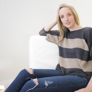 May include: A grey and beige striped sweater with long sleeves. The sweater is worn with blue jeans with distressed details. The person is seated on a white chair.
