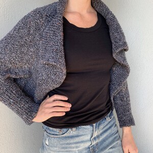 May include: A gray and white speckled knit cardigan with a draped neckline. The cardigan is cropped and has long sleeves.