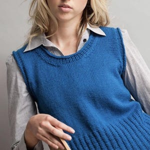 Può includere: Un gilet di maglia blu senza maniche con scollo rotondo. Il gilet ha un orlo a coste ed è indossato sopra una camicia a righe con bottoni.