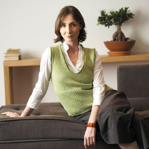 以下が含まれることがあります： 白いシャツと緑のニットベストを着た女性が茶色のソファに座っています。彼女は手首に茶色のブレスレットをしています。