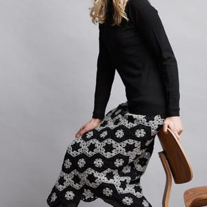 Op de afbeelding: Een vrouw draagt een zwarte turtleneck trui en een grijze gebreide beanie, gecombineerd met een zwart-witte gebreide rok. Ze draagt zwarte laarzen tot aan de knie en zit op een houten stoel.