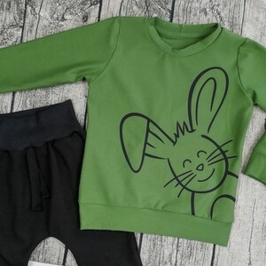 Puede incluir: Una camiseta verde de manga larga con un gráfico de conejo negro. La camiseta se combina con pantalones negros.