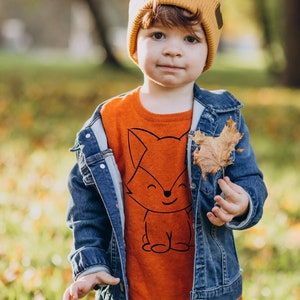 May include: A young person wearing a bright orange sweater with a black fox graphic and a denim jacket. The sweater has a crew neck and the fox graphic is centered on the front. The person is also wearing a yellow beanie with a brown brim.