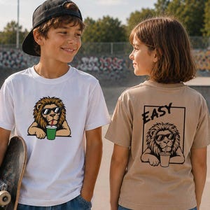 May include: A white t-shirt featuring a graphic of a lion wearing sunglasses and drinking from a green cup, and a tan t-shirt with a black line art graphic of a lion wearing sunglasses, drinking from a cup, and the word EASY inside a rectangle.