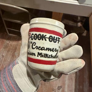 May include: A cylindrical ceramic cup with a cream-colored base, featuring two horizontal red stripes and black text that reads "COOK OUT", "Creamery", and "mium Milkshake". The cup has a smooth, glazed finish and a simple, straight-sided design.