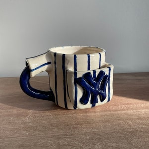 Könnte beinhalten: Eine weiße Keramiktasse mit blauen Streifen und einem blauen New York Yankees Logo an der Seite. Die Tasse hat einen Henkel und eine Tasche an der Seite.