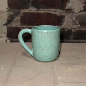 May include: A speckled, light teal ceramic mug with a curved handle. The mug is cylindrical and sits on a light-colored surface, with a brick wall in the background. The mug is designed for hot beverages.