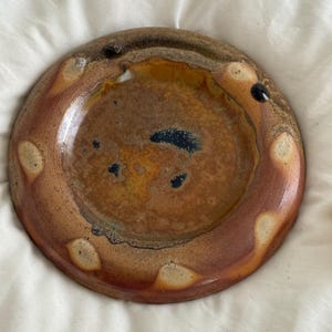 May include: A round ceramic dish with a glossy finish, featuring a mottled brown and amber glaze. The rim is decorated with a series of light beige, oval-shaped spots, while the center displays dark blue and black speckles within the glaze.