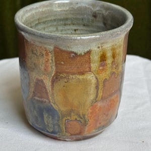 May include: A cylindrical ceramic cup featuring a mottled glaze with overlapping patches of earthy brown, ochre, muted blue, and cream. The rim has a light grey crackle finish, and the exterior surface displays a varied, multi-tonal color palette.
