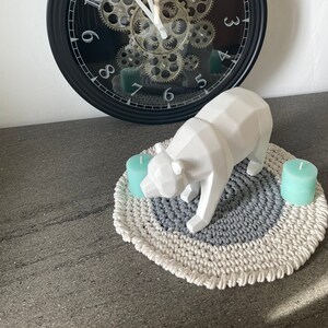 Puede incluir: Una figurita de oso geom&eacute;trico blanco se encuentra sobre una alfombra redonda de crochet gris y blanco con dos velas color turquesa. Un reloj negro con engranajes y n&uacute;meros romanos est&aacute; en el fondo.