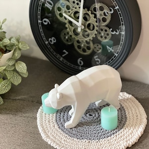 Puede incluir: Una escultura de oso de cer&aacute;mica blanca sobre una alfombra redonda de crochet gris y blanco con dos velas de color verde azulado.