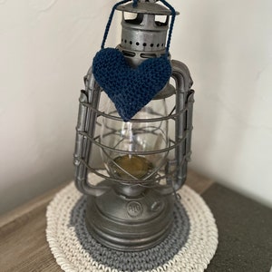 Puede incluir: Una linterna de metal vintage con un globo de vidrio y un coraz&oacute;n azul tejido a crochet colgando de la parte superior. La linterna est&aacute; sentada sobre un posavasos tejido a crochet blanco y gris.
