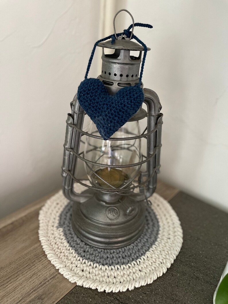 Puede incluir: Una linterna de metal vintage con un globo de vidrio y un coraz&oacute;n azul tejido a crochet colgando de la parte superior. La linterna est&aacute; sentada sobre un posavasos tejido a crochet blanco y gris.