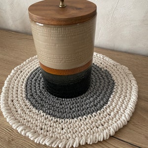 Puede incluir: Un tarro de cer&aacute;mica beige con tapa de madera y pomo de lat&oacute;n, sentado sobre un posavasos redondo de crochet tricolor con hilo blanco, gris y beige.