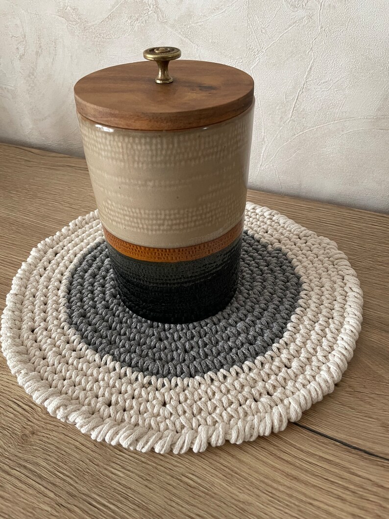 Puede incluir: Un tarro de cer&aacute;mica beige con tapa de madera y pomo de lat&oacute;n, sentado sobre un posavasos redondo de crochet tricolor con hilo blanco, gris y beige.