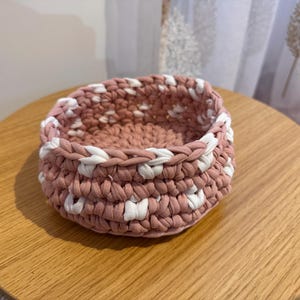 May include: A round, handmade crochet basket in a soft pink colour with white accents. The basket is on a wooden table. The basket is likely used for storage or decoration.