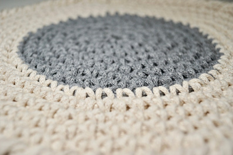 Puede incluir: Una alfombra redonda de crochet con un centro gris y un borde exterior crema.
