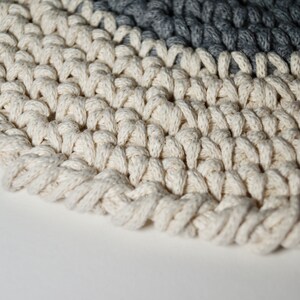 Puede incluir: Primer plano de una alfombra de crochet blanca con un borde de crochet gris. La alfombra est&aacute; hecha de hilo grueso y texturizado y tiene una forma circular.