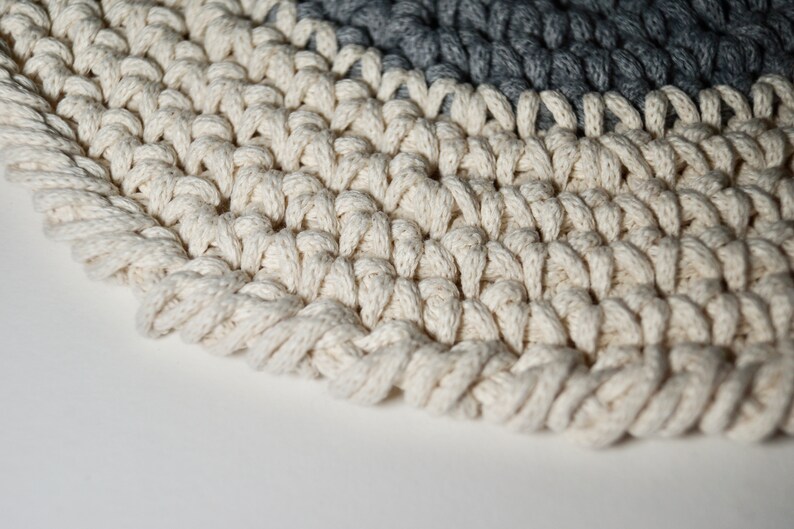 Puede incluir: Primer plano de una alfombra de crochet blanca con un borde de crochet gris. La alfombra est&aacute; hecha de hilo grueso y texturizado y tiene una forma circular.