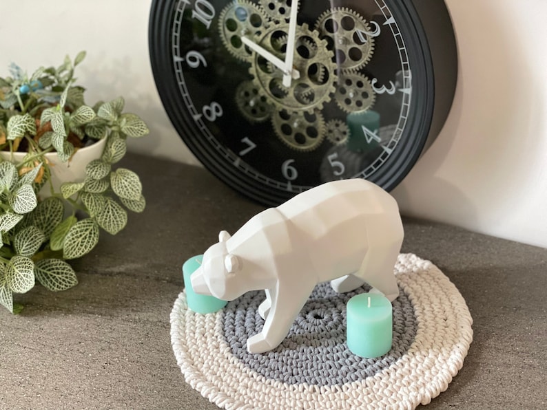 Puede incluir: Una escultura de oso de cer&aacute;mica blanca sobre una alfombra redonda de crochet gris y blanco con dos velas de color verde azulado.