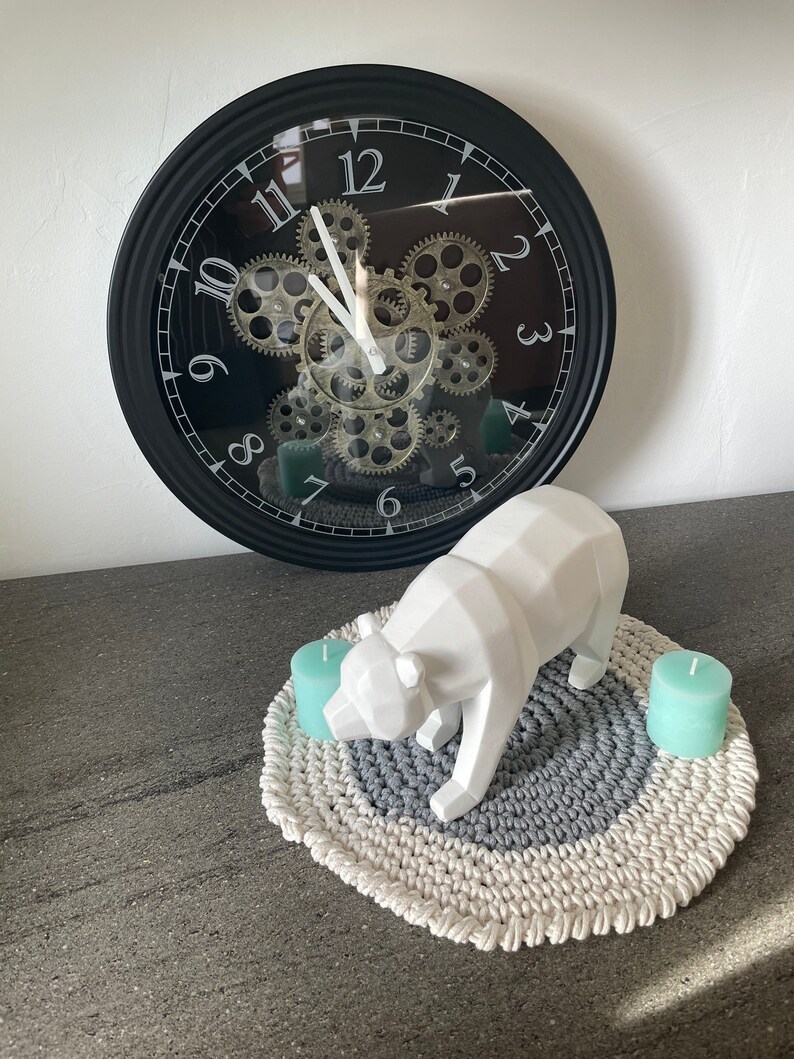 Puede incluir: Una figurita de oso geom&eacute;trico blanco se encuentra sobre una alfombra redonda de crochet gris y blanco con dos velas color turquesa. Un reloj negro con engranajes y n&uacute;meros romanos est&aacute; en el fondo.