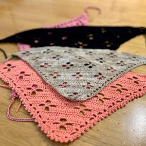 Puede incluir: Tres bufandas triangulares de crochet en rosa, beige y negro. Las bufandas están superpuestas, con la bufanda rosa en la parte inferior y la bufanda negra en la parte superior.