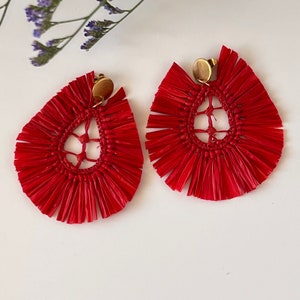 Peut inclure: Une paire de boucles d'oreilles en raphia rouge vif. Chaque boucle d'oreille a une forme de goutte d'eau avec un centre tissé et une frange rayonnante. Les boucles d'oreilles ont un fermoir doré. Le fond est blanc avec une brindille de fleurs violettes.