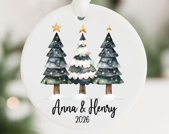 May include: A round white ceramic ornament hanging from a white ribbon. The design features three watercolor-style pine trees with gold stars on top, set against a snowy base. The text Anna & Henry and the year 2026 are printed in black below.