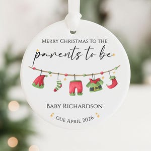 May include: A round white Christmas ornament with a ribbon. It displays the text "MERRY CHRISTMAS TO THE parents to be" and a garland of baby clothes. The bottom text reads "BABY RICHARDSON DUE APRIL 2026". A festive keepsake.