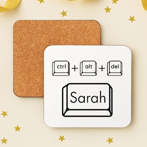 May include: Square white coaster with black keyboard key graphics: ctrl, alt, and del, with the name "Sarah" in a key outline. A cork coaster is next to it. Gold ribbon and star decorations are in the background.