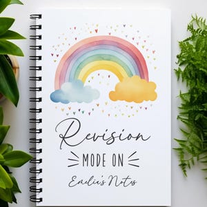 May include: A white spiral-bound notebook featuring a watercolour rainbow illustration with blue and yellow clouds, small colourful hearts, and the black printed text "Revision MODE ON" followed by the handwritten-style text "Emilie's Notes".