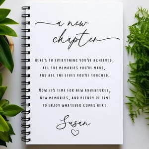 May include: A white spiral-bound notebook featuring the black printed text A new chapter, followed by a message about achievements, memories, and new adventures, ending with the name Susan and a small heart icon centred at the bottom of the page.