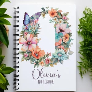 May include: A white spiral-bound notebook featuring a large letter O formed by colourful watercolour-style flowers, leaves, and a blue butterfly. Below the letter, the text reads Olivia's NOTEBOOK in a combination of script and sans-serif fonts.