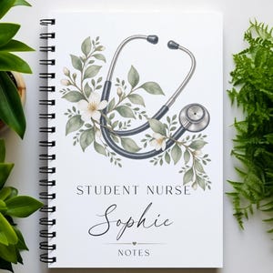 May include: A spiral-bound notebook with a white cover featuring a graphic of a stethoscope intertwined with green leaves and white flowers. The text on the cover reads STUDENT NURSE, the name Sophie in a script font, and the word NOTES below.