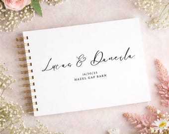 以下が含まれることがあります： 金色のリング綴じの白いゲストブック。表紙には「Lucas & Daneila」の名前が筆記体で書かれ、日付「16/10/25」と「HAZEL GAP BARN」も記載されています。ピンクと白の花に囲まれています。