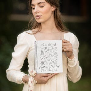 May include: A white spiral-bound notebook with the text "Getting Hitched Future Mrs. Smith" and wedding-themed doodles. The notebook is held in front of a person wearing a cream-colored dress.