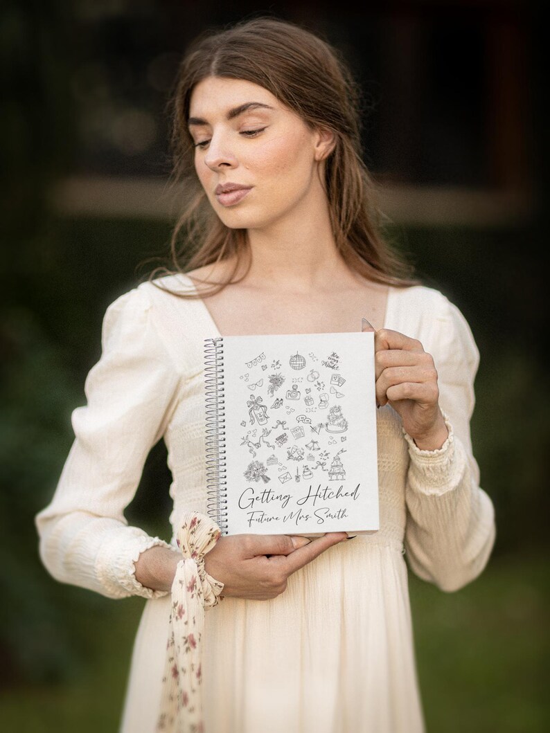 May include: A white spiral-bound notebook with the text "Getting Hitched Future Mrs. Smith" and wedding-themed doodles. The notebook is held in front of a person wearing a cream-colored dress.