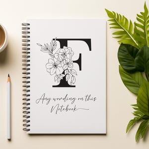 May include: A white spiral-bound notebook featuring a black floral design and the letter "F". The notebook has the text "Any wording on this Notebook". A cup of coffee and a pencil are next to the notebook, with green leaves in the background.