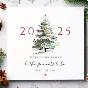 Può includere: Un biglietto di Natale con un'illustrazione ad acquerello di un albero di Natale. Il biglietto recita "2025" e "Buon Natale ai futuri genitori" con i nomi "Matt & Liv" sotto. Il biglietto ha un cuore rosso in basso.