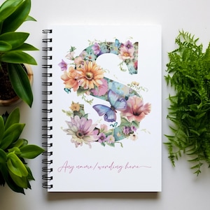 May include: A white spiral-bound notebook with a floral "S" design, featuring butterflies and flowers in pink, orange, and purple hues. The text "Any name/wording here" is written in cursive. Green plants are arranged around the notebook.