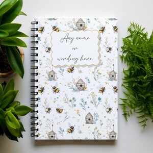May include: A patterned notebook with a bee and beehive design. The cover includes the text "Any name or wording here" within a decorative frame. Green plants are arranged around the notebook.